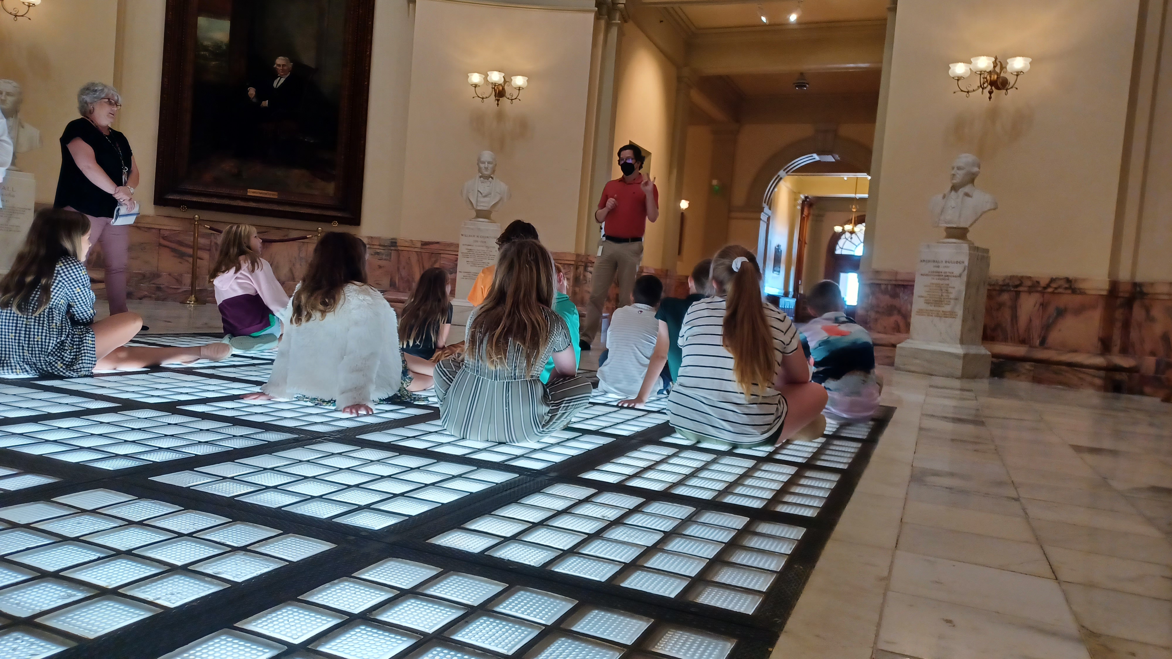 Museum Educator stands at the front of a group of seated children, leading their tour of the Georgia Capitol Museum. 