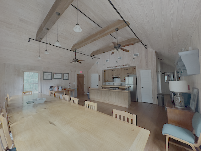 A dining room with long wood table and six chairs on each side  with a kitchen area in the background.