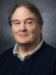Man with brown hair wearing grey shirt and navy pullover sweater