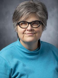 Woman with short hair wearing glasses and an aqua turtleneck.