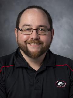 Bearded man with glasses wearing a black golf shirt with the the UGA "Big G" logo on it.