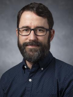 Man with a beard and glasses wearing a patterned blue button down shirt.