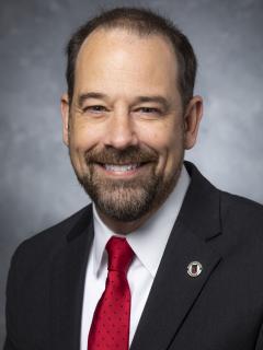 Man with short dark hair and a beard wearing a dark suit, white shirt, and red tie. On his lapel is a pin bearing the Libraries logo.
