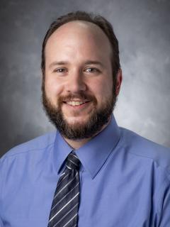 Man with dark hair and beard wearing a blue button up shirt and diagonally striped tie.
